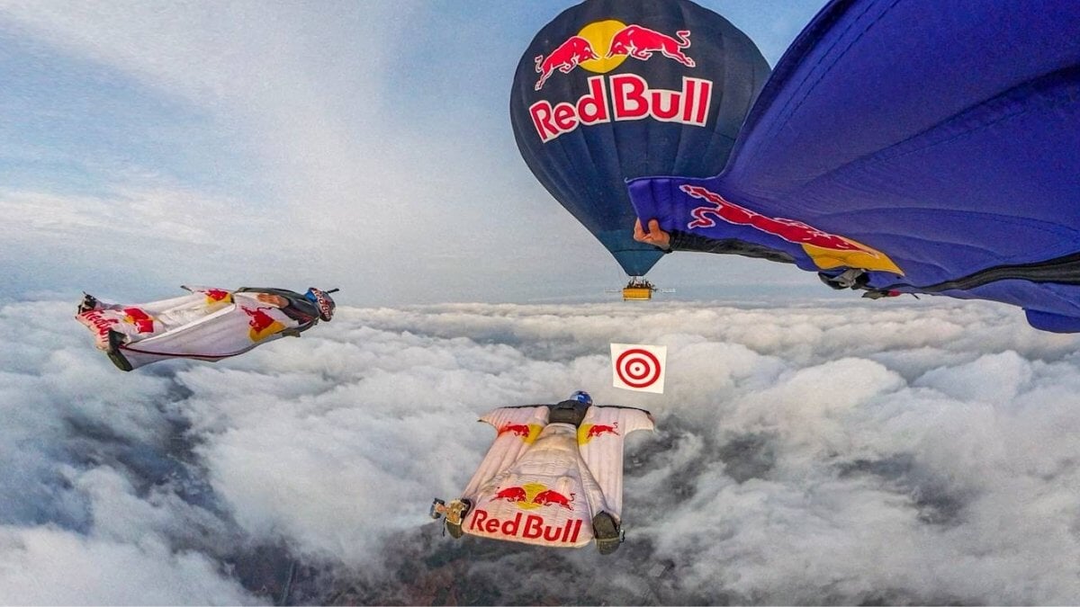 Photo aérienne extrême : pilotes en combinaisons blanches sur ailes volantes Red Bull au-dessus des nuages, avec ballon bleu Red Bull et cible blanche.